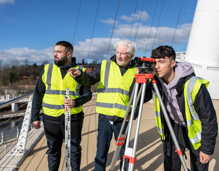 Three individuals wearing high-visibility vests conducting a land surveying exercise on a bridge. One is looking through a theodolite, while another holds a measuring staff, and a lecturer observes under a bright blue sky. Three individuals wearing high-visibility vests conducting a land surveying exercise on a bridge. One is looking through a theodolite, while another holds a measuring staff, and a lecturer observes under a bright blue sky.